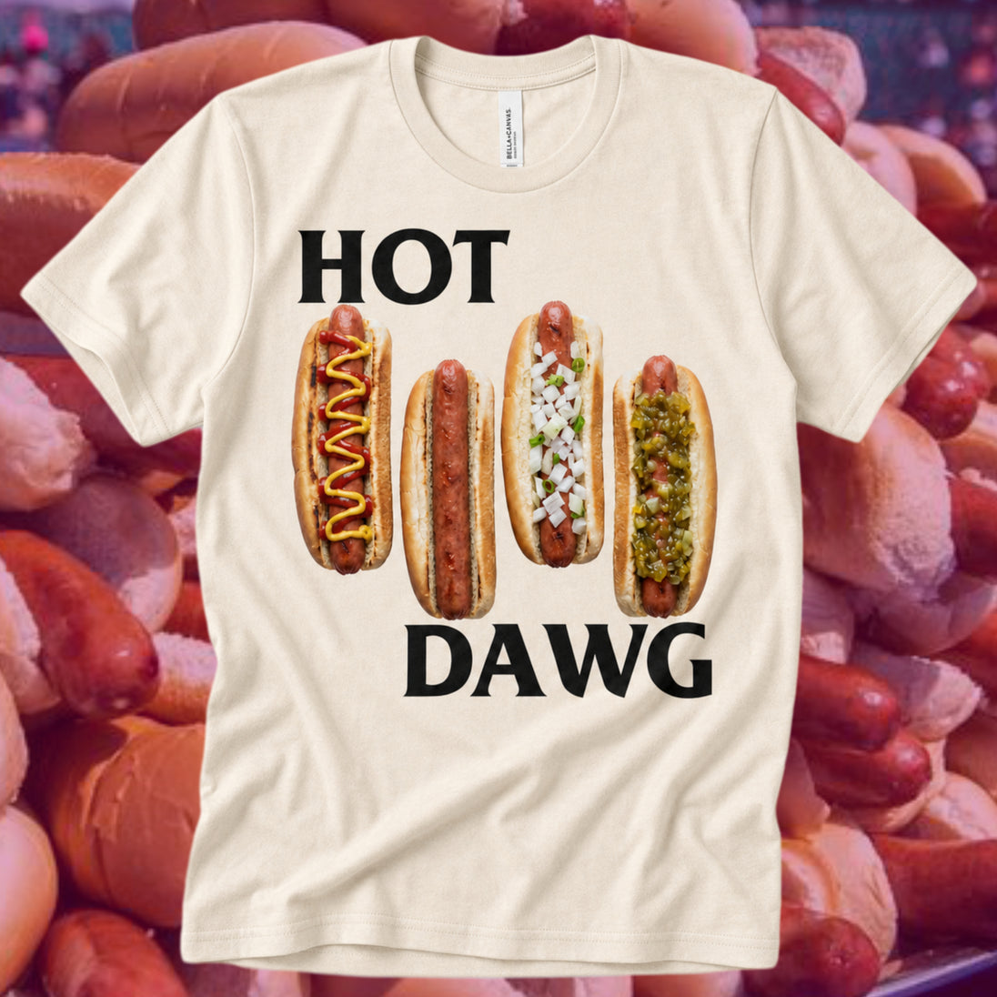 A cream-colored t-shirt with a humorous graphic that reads 'HOT DAWG' in bold black letters, featuring four illustrated hot dogs with various toppings. The shirt is displayed against a background of real hot dogs.
