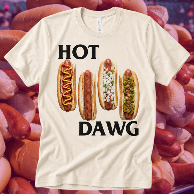 A cream-colored t-shirt with a humorous graphic that reads 'HOT DAWG' in bold black letters, featuring four illustrated hot dogs with various toppings. The shirt is displayed against a background of real hot dogs.