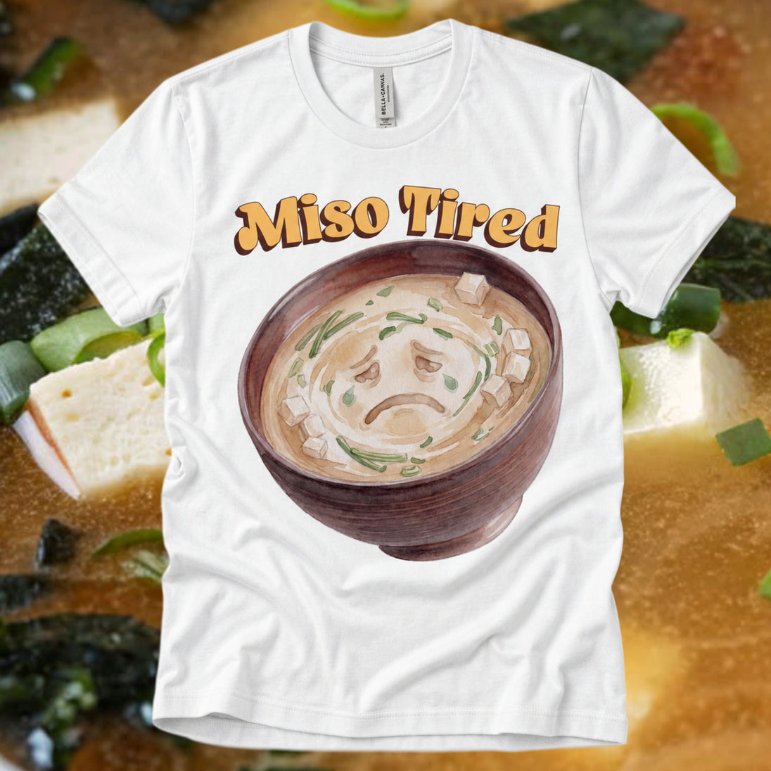 A white graphic t-shirt featuring a sad face illustration of miso soup in a bowl with the text 'Miso Tired' above it, displayed against a background of miso soup ingredients.