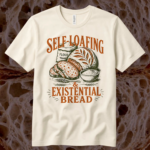 An off-white short-sleeve t-shirt with a retro-style graphic of bread, a flour sack, and wheat, featuring the text 'SELF LOAFING & EXISTENTIAL BREAD' in orange-brown. The shirt is displayed flat against a textured, organic brown background.