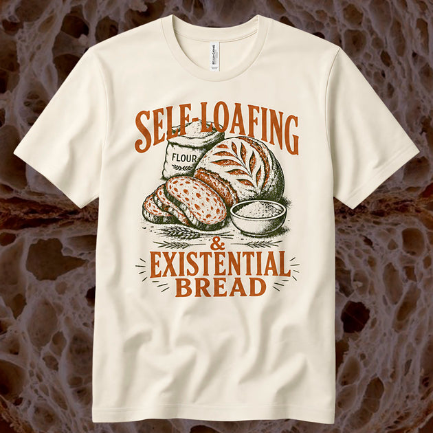An off-white short-sleeve t-shirt with a retro-style graphic of bread, a flour sack, and wheat, featuring the text 'SELF LOAFING & EXISTENTIAL BREAD' in orange-brown. The shirt is displayed flat against a textured, organic brown background.
